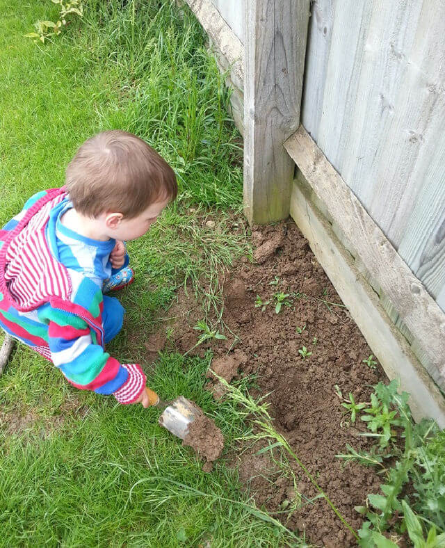 toddler in stripy bright jumper digging in muddy patch next to a fence