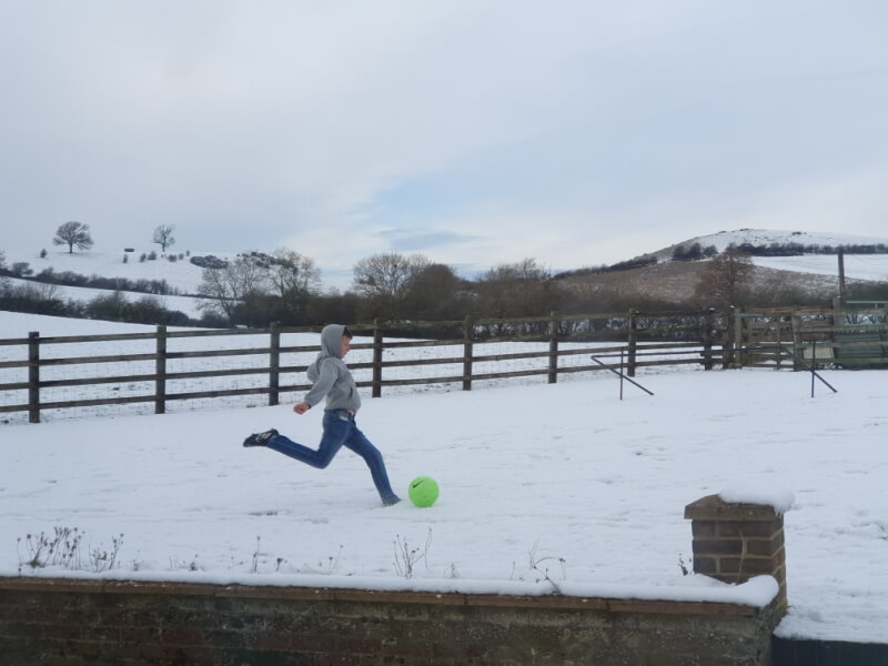 boy about to kick a football in snowy garden wearing blue jeans and grey hoody