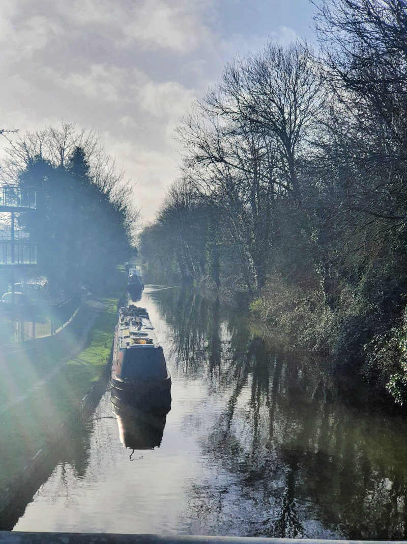 looking down the canal while the morning sun shines on it and a canal boat moored on the left with trees